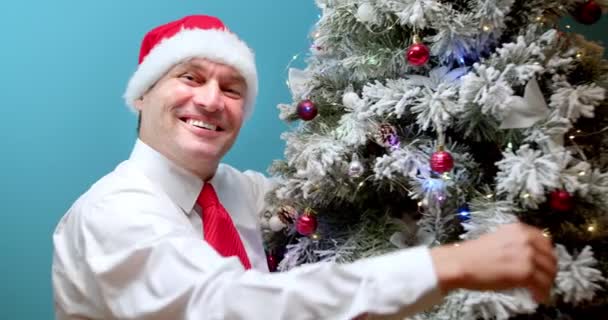 圣诞前夕 一个头戴圣诞礼帽 头戴圣诞礼帽 头戴圣诞礼帽 面带微笑的男人看着摄像机 从快乐中拥抱着圣诞树 — 图库视频影像