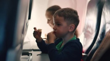 Küçük çocuk uçakta ailesiyle uçarken hamburger yiyor ve yemeğin tadını çıkarıyor. Ailesi olan çocuklar uçakta uçar, çocukluk yolculuğu konsepti.