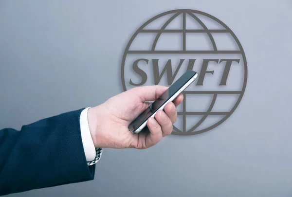 Takım elbiseli bir iş adamı, Mart 04 'te Moskova' da bir duvarda SWIFT logosu önünde akıllı telefon tutuyor. Rusya 'da 2022