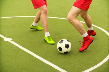 Oyuncular sahada mini futbol oynarlar. Bacaklar, spor ayakkabılar ve bir top. Yeşil futbol sahası.