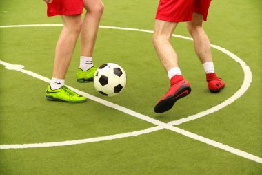 Oyuncular sahada mini futbol oynarlar. Bacaklar, spor ayakkabılar ve bir top. Yeşil futbol sahası.