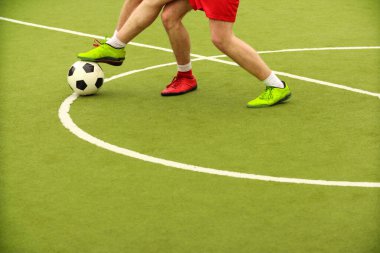 Oyuncular sahada mini futbol oynarlar. Bacaklar, spor ayakkabılar ve bir top. Yeşil futbol sahası.