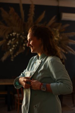 Kadın aşçı işe hazırlanıyor. Önlük giyer, düğmelerini bağlar ve kollarını sıvar.