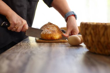 Parlak bir köy mutfağında kadın şef kahvaltı hazırlıyor. Ekmeği keser ve peynirle yayar. Çiçekli mutfak masası ve mutfak tahtası, ekmek, domates, yeşillik