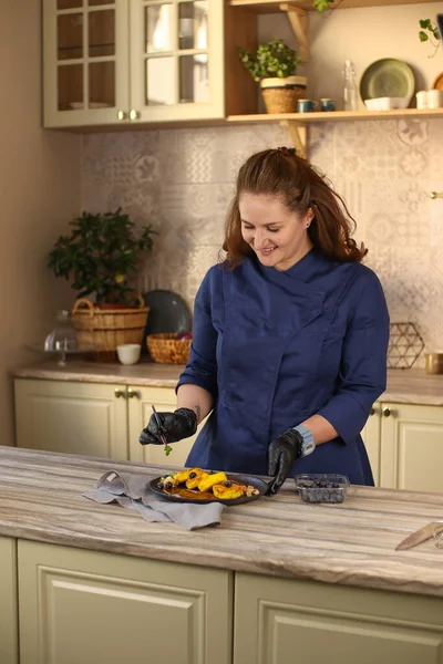 Female chef in a bright country kitchen.Dressed in a blue apron and black pants. Makes cheesecakes with salted caramel and fruit