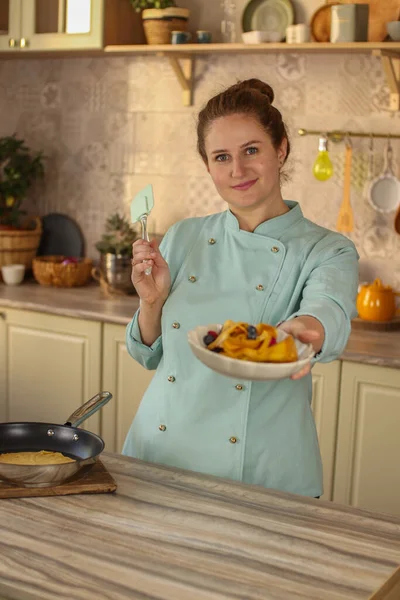 Female chef prepares pancakes in a bright kitchen in a rustic style. Mint coat apron. Homemade pancakes with berries. Chef at home