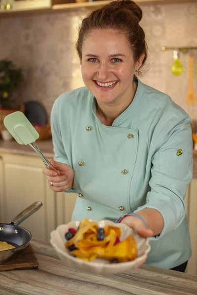 Female chef prepares pancakes in a bright kitchen in a rustic style. Mint coat apron. Homemade pancakes with berries. Chef at home