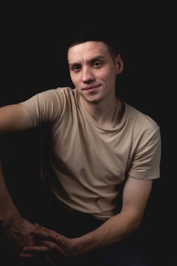 Siyah arka planda poz veren yakışıklı bir genç. Stüdyo fotoğrafı ve tek bir ışık kaynağı. Gülümseyen adam neşeli bir şekilde stüdyoda poz veriyor. Ciddi bir adam dirseği dizinde oturur..