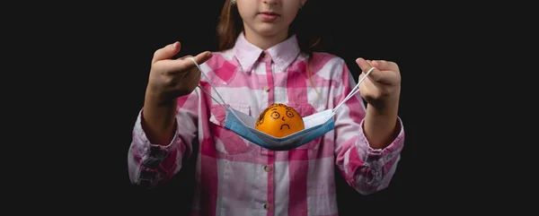 一个小女孩在她面前拿着一个蓝色的医疗面具 上面有一条橘红色的线 用黑色毛笔画的普通话就像一个没有面具的人 用一个光源隔离在黑色背景上的演播室肖像 — 图库照片