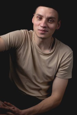 Siyah arka planda poz veren yakışıklı bir genç. Stüdyo fotoğrafı ve tek bir ışık kaynağı. Gülümseyen adam neşeli bir şekilde stüdyoda poz veriyor. Neşeli bir adam dirseği dizinde oturur..