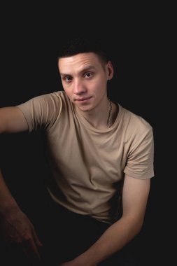 Siyah arka planda poz veren yakışıklı bir genç. Stüdyo fotoğrafı ve tek bir ışık kaynağı. Gülümseyen adam neşeli bir şekilde stüdyoda poz veriyor. Ciddi bir adam dirseği dizinde oturur..