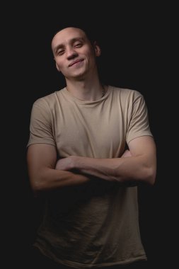 Siyah arka planda poz veren yakışıklı bir genç. Stüdyo fotoğrafı ve tek bir ışık kaynağı. Kendine güvenen adam kollarını önünde kavuşturur ve gülümser..