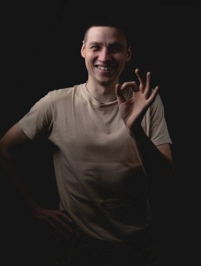 Siyah arka planda poz veren yakışıklı bir genç. Stüdyo fotoğrafı ve tek bir ışık kaynağı. Gülümseyen adam neşeli bir şekilde stüdyoda poz veriyor. Gülen adam sol eliyle işaret veriyor..