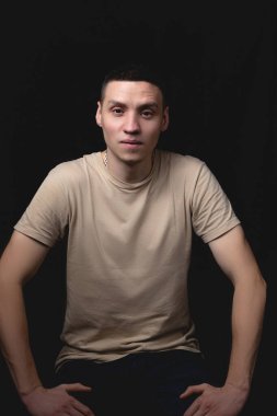 Siyah arka planda poz veren yakışıklı bir genç. Stüdyo fotoğrafı ve tek bir ışık kaynağı. Gülümseyen adam neşeli bir şekilde stüdyoda poz veriyor. Adam dizlerinin üstüne çökmüş oturuyor..