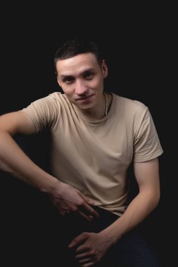 Siyah arka planda poz veren yakışıklı bir genç. Stüdyo fotoğrafı ve tek bir ışık kaynağı. Gülümseyen adam neşeli bir şekilde stüdyoda poz veriyor. Neşeli bir adam dirseği dizinde oturur..
