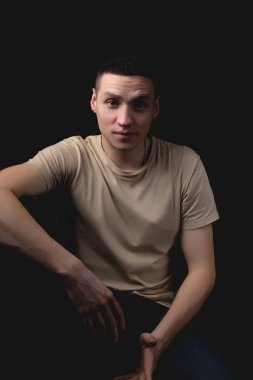 Siyah arka planda poz veren yakışıklı bir genç. Stüdyo fotoğrafı ve tek bir ışık kaynağı. Gülümseyen adam neşeli bir şekilde stüdyoda poz veriyor. Arkadaş canlısı bakışlı bir adam elleri dizinde oturur..