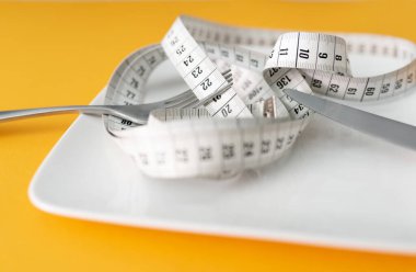 tape measure on plate with fork and knife against orange table background, dieting and loosing weight concept