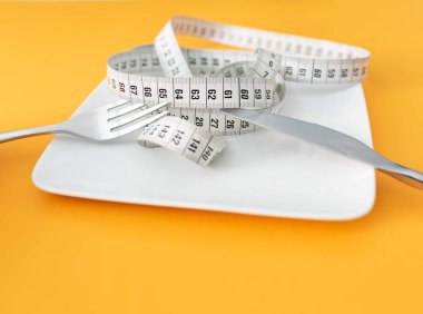 tape measure on plate with fork and knife against orange table background with copy space, dieting and loosing weight concept