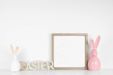 Beyaz bir rafta Paskalya süsleriyle ahşap çerçeve yap. Modern cam tavşan ve tahta Paskalya tabelası. Beyaz bir duvara karşı kare çerçeve. Boşluğu kopyala.