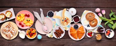 Eğlenceli Paskalya kahvaltısı ya da brunch masası sahnesi. Koyu ahşap bir arkaplanda manzaranın üstünde. Tavşan gözlemesi, yumurta yuvası, civciv meyvesi ve ilkbahar yiyecekleri seçimi..