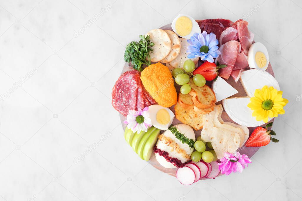 Spring or Easter theme charcuterie board against a white marble background. Variety of cheese, meat, fruit and vegetable appetizers. Top view.
