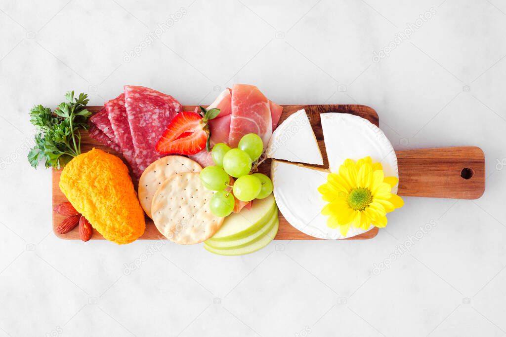 Spring or Easter theme charcuterie board against a white marble background. Selection of cheese, meat, fruit and vegetable appetizers. Above view.