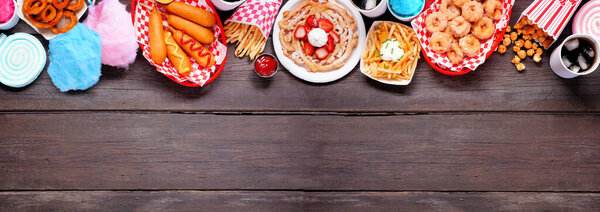 Carnival theme food top border over a dark wood banner background. Overhead view with copy space. Summer fair concept. Corn dogs, funnel cake, cotton candy and snacks.