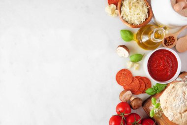 Pizza making ingredients including cheese, pepperoni, tomatoes and basil. Above view side border against a white marble background. Copy space.