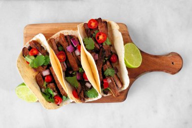 Tacos with steak, tomatoes, onion and cilantro. Overhead view serving board on a white marble background.