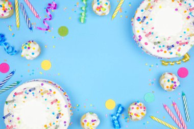 Birthday party frame on a blue background. Overhead view with cakes, party hats and confetti. Copy space.