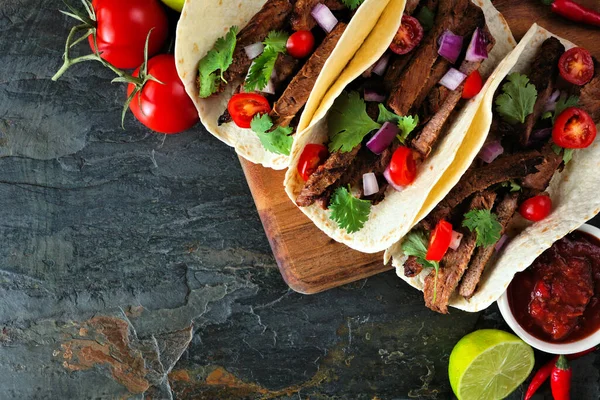 Mexican tacos with steak, tomatoes, onion and cilantro. Top down close up on a dark background with copy space.