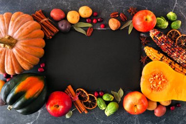 Fall food frame of pumpkins, apples, squash and an assortment of vegetables surrounding a blank slate serving board. Top down view on a dark stone background with copy space.
