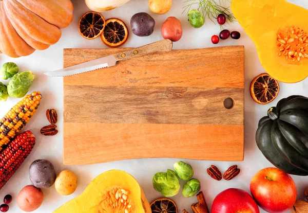 Autumn food frame of pumpkins, apples, squash and assorted vegetables surrounding a blank wood serving board. Overhead view on a white marble background with copy space.