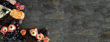Halloween theme charcuterie corner border against a dark stone banner background. Assorted cheeses and meat appetizers. Copy space.