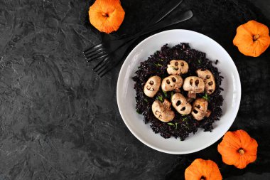 Halloween risotto with skull mushrooms and black rice. Above view over a dark black background. Copy space.