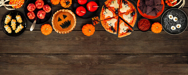 Fun Halloween dinner party top border over a dark wood banner background. Overhead view. Pizza, jack o lantern pumpkin pie, candy apples, eyeball spaghetti, snacks and spooky punch.