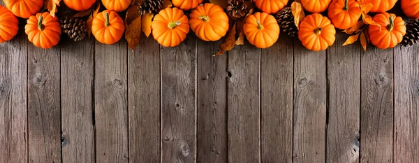Fall top border of pumpkins, leaves and pine cones. Overhead view on a rustic dark wood banner background with copy space.