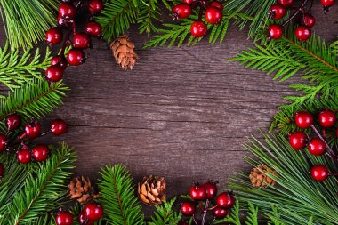 Her zaman yeşil dalları, kırmızı böğürtlenleri ve çam kozalakları olan bir Noel çerçevesi. Koyu ahşap arkaplan üzerindeki üstten aşağı görünüm.