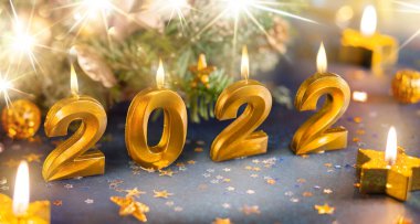 2022 yılbaşı tebrik kartı, mumlardan, ışıltılı konfetilerden ve Noel ağacı çelenklerinden yapılma mavi arka planda şenlik süsleri olan altın sayılar..