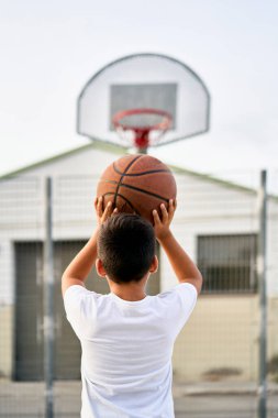 Tatlı genç bir çocuk yaz mevsiminde sokak bahçesinde basketbol oynar. Dışarıda turuncu basketbol topu olan beyaz tişörtlü bir genç. Hobi, aktif yaşam tarzı, çocuklar için spor aktiviteleri.