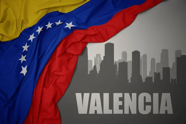 バレンシアという文字のある都市の抽象的なシルエット灰色の背景にベネズエラのカラフルな国旗を振って近く — ストック写真