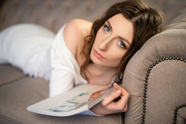 Bella ragazza in abito bianco è sdraiata sul divano con una rivista di moda — Foto Stock