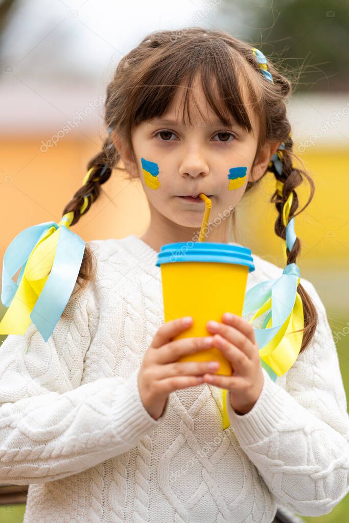 A little Ukrainian girl with two pigtails and yellow and blue ribbons drinks a hot drink from a glass of patriotic colors like the Ukrainian flag. Stop the war in Ukraine. Volunteer Help for Refugees. High quality photo