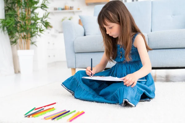 A cute little girl sits on the floor at home and draws with colored pencils. Child development and creativity for preschoolers and younger students. High quality photo