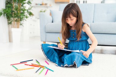 Sevimli küçük bir kız evde yerde oturuyor ve renkli kalemlerle resim çiziyor. Anaokulu öğrencileri ve daha genç öğrenciler için çocuk gelişimi ve yaratıcılık. Yüksek kalite fotoğraf