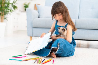 Kot elbiseli, saçına perçem takan sevimli bir kız halının üzerindeki bir odada uzanıyor ve renkli kalemlerle resim çiziyor. Yanında dört bacaklı bir arkadaş var. Küçük, oynak, tüylü bir kedi yavrusu. Yüksek kalite fotoğraf