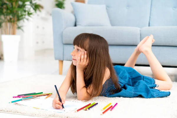 Cute funny girl in a denim sundress and bangs in her hair lies in the room on the carpet on the warm floor and draws with colored pencils. Homeschooling and creative development of the child. High quality photo