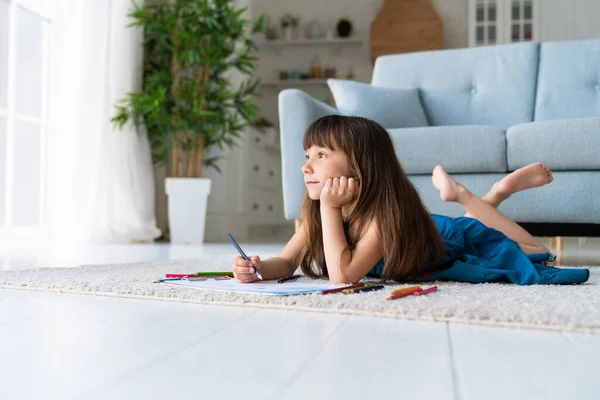 Cute funny girl in a denim sundress and bangs in her hair lies in the room on the carpet on the warm floor and draws with colored pencils. Homeschooling and creative development of the child. High quality photo