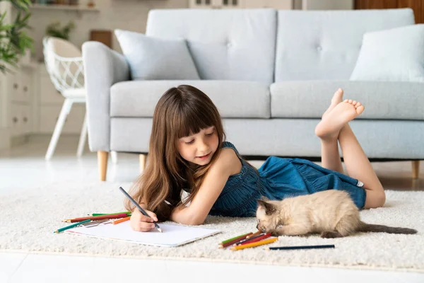 デニムのサンドレスと前髪のある可愛い面白い女の子は カーペットの上の部屋にあり 色鉛筆で描かれています 彼女の横にある4つの足の友人 小さな遊び心のあるふわふわの子猫です 高品質の写真 — ストック写真