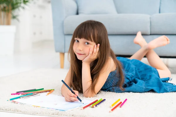 드레스 머리에 송곳니를 귀여운 소녀가 바닥에 색연필로 그림을 그린다 과정이다 스톡 이미지